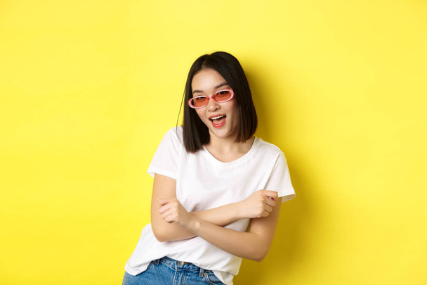 Concepto de moda y estilo de vida. Mujer asiática atrevida y confiada en gafas de sol de moda con aspecto seguro de sí mismo en la cámara, de pie sobre el fondo amarillo - Foto, Imagen