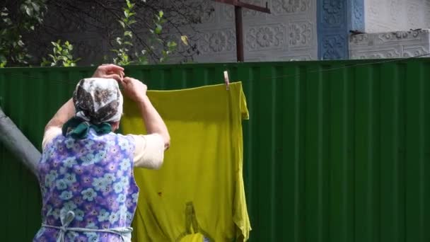Mujer mayor rural de pie atrás y colgando la ropa en pinzas de ropa al aire libre - Imágenes, Vídeo