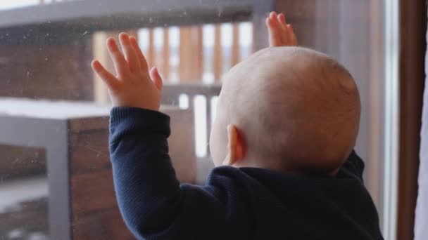 Un niño lindo mira por la ventana y golpea el cristal con las manos - Imágenes, Vídeo