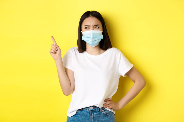 Covid, salud y concepto pandémico. Mujer asiática con máscara médica y camiseta blanca apuntando con el dedo al logotipo de la esquina superior izquierda, mostrando promoción, fondo amarillo - Foto, imagen