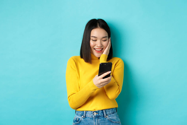 Image de femme asiatique heureuse lisant le message sur l'écran de téléphone mobile et souriant, chat dans l'application smartphone, debout sur fond bleu - Photo, image