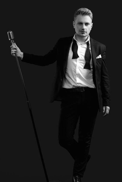 Portrét pohledné blond muž zpěvák v elegantním smokingu a motýlek pózování s vintage mikrofon na černém pozadí studia - Fotografie, Obrázek