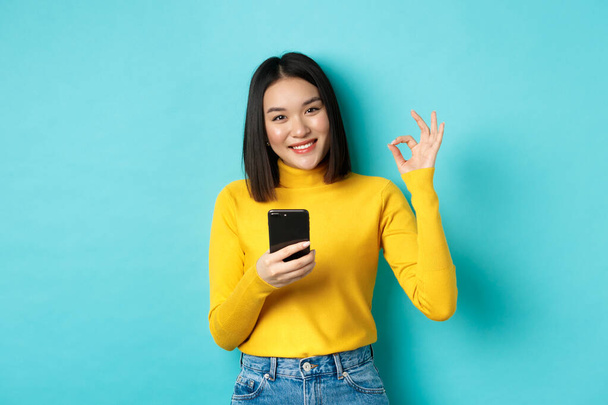 Concepto de comercio electrónico y compras en línea. Retrato de mujer asiática mostrando signo OK y el uso de teléfono móvil, aplicación de alabanza, de pie sobre fondo azul - Foto, imagen