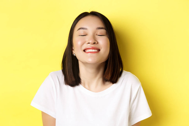 Zblízka šťastný a uvolněný asijské žena těší slunce, usmívá se se zavřenýma očima a vypadá radostně, stojí nad žlutým pozadím - Fotografie, Obrázek