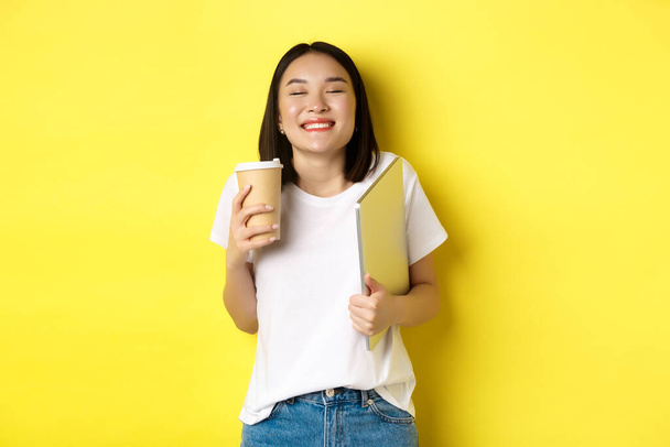 Boldog fiatal nő élvezi kávé kávézó és mosolygós, gazdaság csésze és laptop, álló sárga háttér - Fotó, kép