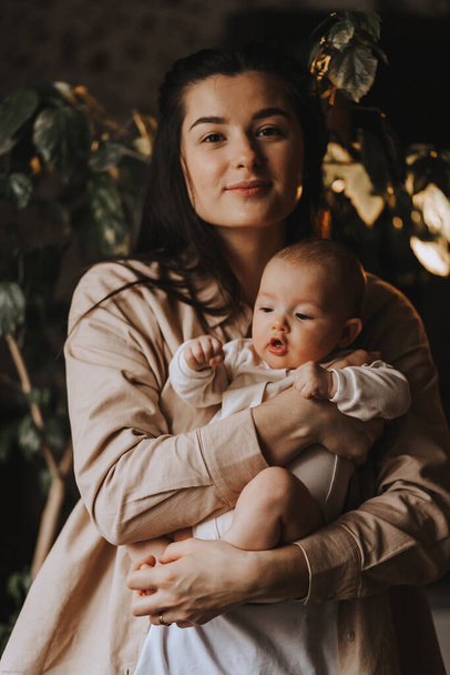 молода брюнетка мама і її новонароджена дитина в руках, домашнє фото в темній кімнаті біля вікна
 - Фото, зображення