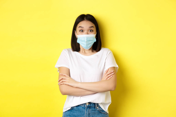 Covid, health care and pandemic concept. Asian woman in white t-shirt and medical mask cross arms on chest and looking surprised at camera - Foto, immagini