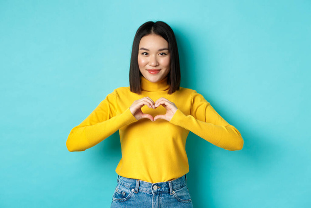 Приваблива японська дівчина в жовтому светрі, показує жест серця і каже, що я люблю тебе, дивлячись щиро на камеру, стоїть над синім тлом
 - Фото, зображення