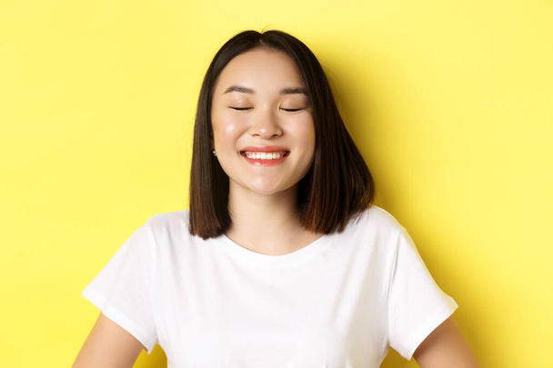 Close up of happy, romantic asian girl dreaming of something, close eyes and smiling delighted, standing over yellow background - Фото, зображення