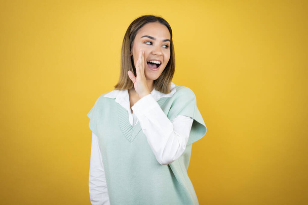 Pretty young woman standing over yellow background with hand on mouth telling secret rumor, whispering malicious talk conversation - Photo, Image