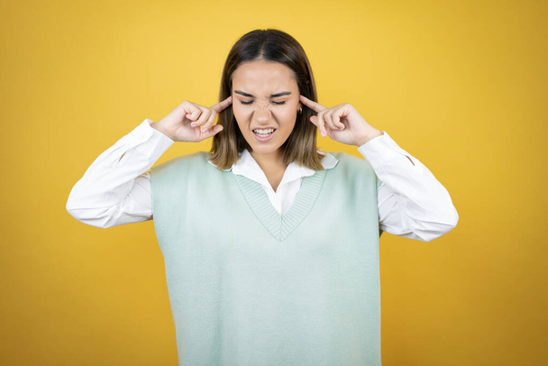 Pretty young woman standing over yellow background covering ears with fingers with annoyed expression for the noise of loud music. Deaf concept. - Photo, Image