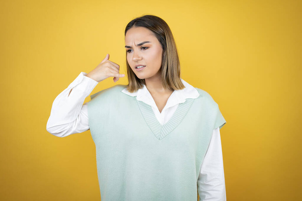 Pretty young woman standing over yellow background doing phone gesture with hand and fingers like talking on the telephone confused - Fotoğraf, Görsel
