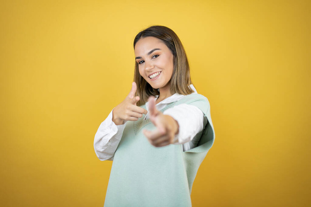 Гарненька молода жінка стоїть над жовтим тлом, вказуючи на вас і камеру з пальцями, посміхаючись позитивно і весело
 - Фото, зображення