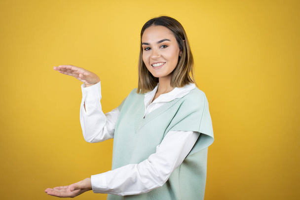 Pretty young woman standing over yellow background gesturing with hands showing big and large size sign, measure symbol. Smiling . Measuring concept. - Photo, Image