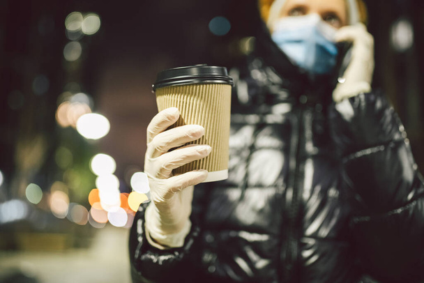European city life due to quarantine regulations during covid19 pandemic. Adult woman resident city observes safety measures, wears face masks and gloves, uses cell phone and holds cup hot beverage. - Photo, Image