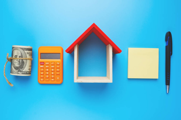 Een foto huis miniatuur, met nep geld, notitieblok, rekenmachine, en pen op blauwe achtergrond. - Foto, afbeelding