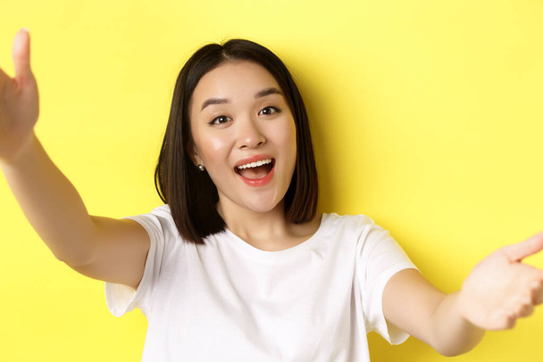 Imagen de una hermosa mujer asiática sosteniendo la cámara del teléfono inteligente y sonriendo, tomando selfie o video chat, de pie sobre fondo amarillo - Foto, Imagen