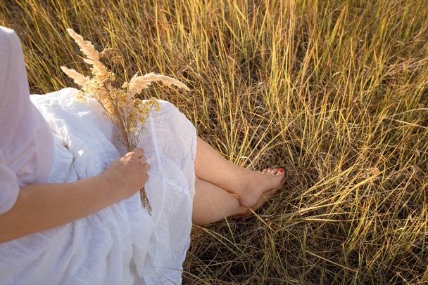 Boso ubrana kobieta siedzi na polu i trzyma bukiet suszonej trawy na kolanach. Przestrzeń kopiowania - Zdjęcie, obraz