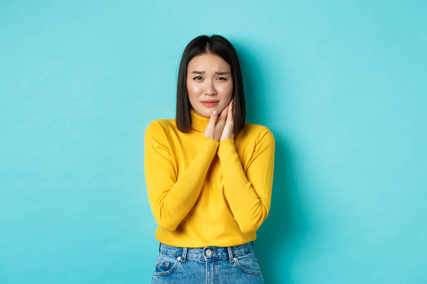 Junge asiatische Frau braucht Zahnarzt, berührt Wange und runzelt die Stirn von schmerzhaften Zahnschmerzen, vor blauem Hintergrund stehend. Gesundheitskonzept - Foto, Bild