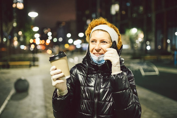 Femme âgée tenant café et téléphone portable, portant un masque médical protecteur et des gants en latex blanc, marchant à l'extérieur, observant la distance sociale par pandémie de coronavirus et la quarantaine en Europe. - Photo, image