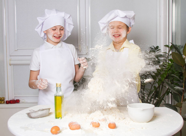 Aşçı gibi giyinmiş iki tatlı Avrupalı çocuk un, süt ve yumurta yoğuruyorlar, çocuklar mutfakta oynuyorlar. - Fotoğraf, Görsel