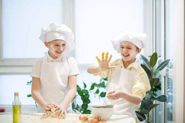 Aşçı gibi giyinmiş iki tatlı Avrupalı çocuk un, süt ve yumurta yoğuruyorlar, çocuklar mutfakta oynuyorlar. - Fotoğraf, Görsel