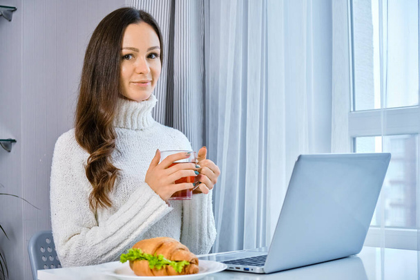 Happy casual beautiful woman working on a laptop sitting at the table in the kitchen and looks at the camera - Foto, imagen