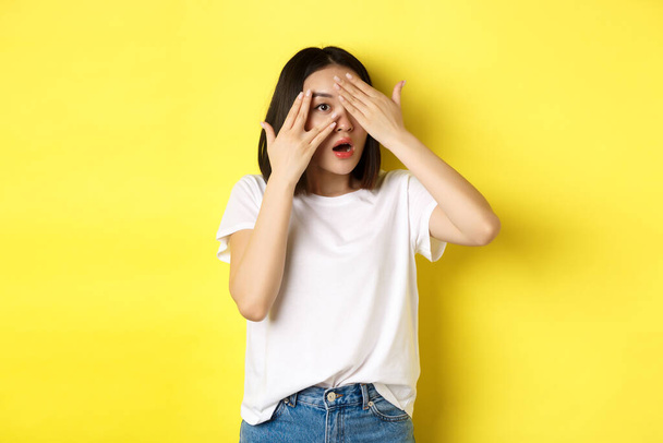 Intrigued asian girl waiting for surprise, peeking through fingers on eyes, smiling happy, standing over yellow background - 写真・画像