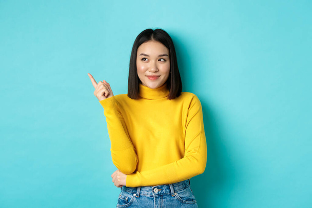 Alışveriş konsepti. Sarı kazaklı şık Asyalı kadın model, gülümseyen ve sol elini gösteren, memnun yüzüyle reklamı gösteren, mavi arka planda duran - Fotoğraf, Görsel