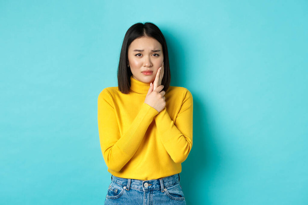 Victim of domestic violance, young asian woman, touching cheek and frowning, being slapped in face, standing over blue background - Фото, зображення