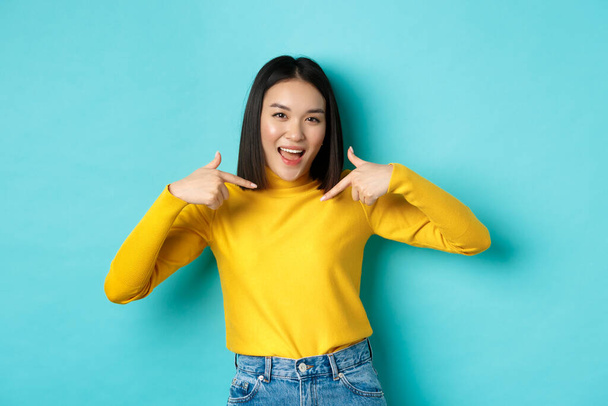 Beauty and fashion concept. Confident and sassy asian woman pointing at herself, smiling cheeky at camera, being professional, standing over blue background - Photo, Image