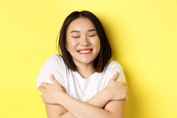 Close up of romantic asian girl hugging herself and dreaming, close eyes and smile while imaging something tender, standing over yellow background - Photo, Image