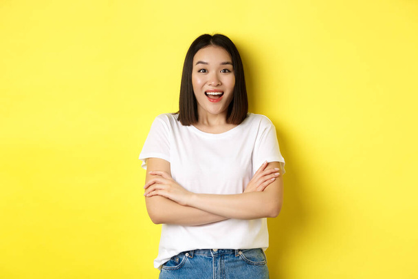 Güzellik ve moda konsepti. Beyaz tişörtlü mutlu Asyalı kadın, göğsünde çapraz kollar, kameraya bakıyor, sarı arka planda duruyor. - Fotoğraf, Görsel