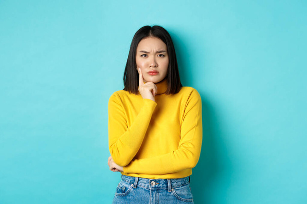 Image of pensive and serious asian woman touching chin, frowning and staring at camera puzzled, making difficult choice, standing over blue background - Фото, зображення