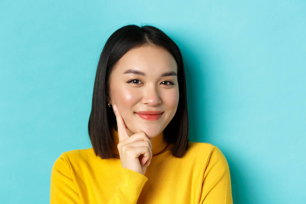 Beauty and makeup concept. Close up of thoughtful asian woman looking pleased at camera and smile, having an idea, standing over blue background - Foto, Imagem