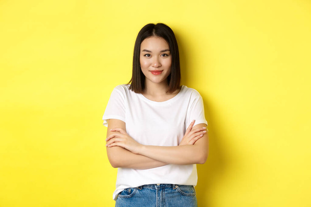 Confident and stylish asian woman cross arms on chest and smiling, standing over yellow background - Фото, изображение