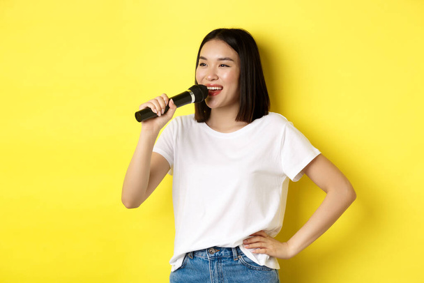 Szczęśliwa azjatycka kobieta śpiewa piosenkę w karaoke, trzymając mikrofon i patrząc na bok z wesołym uśmiechem, stojąc nad żółtym tle - Zdjęcie, obraz