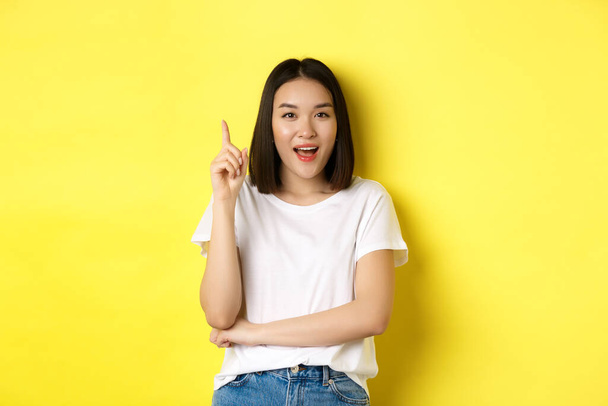 美しさとファッションのコンセプト。興奮したアジアの少女は、ユーレカジェスチャーで指を上げます,アイデアと笑顔をピッチング,黄色の背景の上に立って - 写真・画像