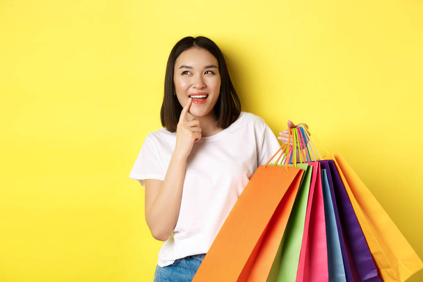 Penzitivní mladá žena se zaujatě usmívá, ukazuje nákupní tašky, přemýšlí o koupi něčeho, stojí nad žlutým pozadím - Fotografie, Obrázek