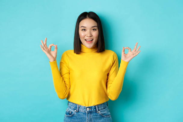 Веселая азиатская модель, показывающая нормальные жесты, улыбающаяся и выглядящая впечатленной, восхваляющая продукт, стоящая на синем фоне - Фото, изображение