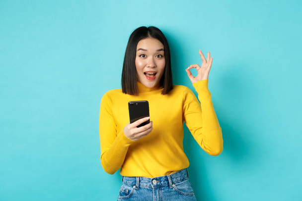 E-commerce and online shopping concept. Portrait of asian woman showing OK sign and using mobile phone, recommend application, standing over blue background - Fotografie, Obrázek