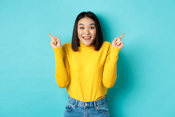 Krása a módní koncept. Veselý asijské dívka ukazuje dvě promo nabídky, ukazuje prsty bokem na levé a pravé reklamy a usmívá se, stojí nad modrým pozadím - Fotografie, Obrázek