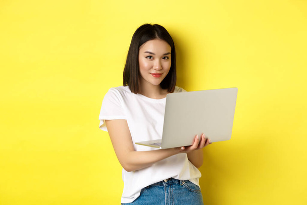 Joven mujer asiática trabajando freelance, utilizando el ordenador portátil y sonriendo, de pie sobre fondo amarillo - Foto, Imagen