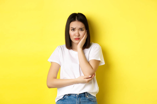 Sad asian woman touching cheek and frowning, having toothache, need see dentist, standing gloomy against yellow background - Photo, Image