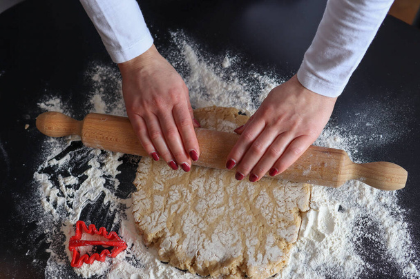 Female hands with a wooden rolling pin roll out the cookie dough on the kitchen table covered with flour. Homemade prepare and decoration food concept - Photo, Image