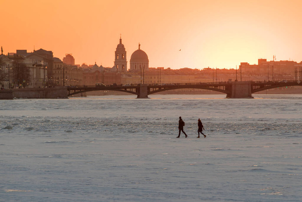 人々は冬の日没でサンクトペテルブルクの凍結ネヴァ川の交差点,橋や教会を背景に.早春に川の氷の上に非常に危険な散歩.冬のフェアウェイに沿って歩く - 写真・画像