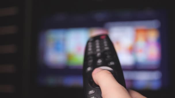 Mans hand selects internet tv channels with remote control, close-up. Person controls TV using a modern remote control. A man watches smart TV and uses black remote control. Blurry tv scrolls pages - Footage, Video
