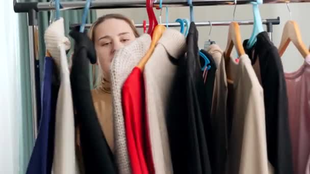 Beautiful smiling woman behind long rack of clothes on hangers choosing dress to wear - Footage, Video