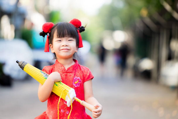 Belle fille asiatique tenant parapluie avec fermé, style vintage jaune. Les enfants sourient doux et heureux. L'enfant portait une robe rouge du Nouvel An chinois et une pince à cheveux rouge fourrure. Adorable enfant de 3-4 ans. - Photo, image
