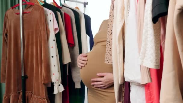 Zwangere jonge vrouw loopt tussen lange rekken van kleding op hangers in winkelcentrum of boetiek - Video